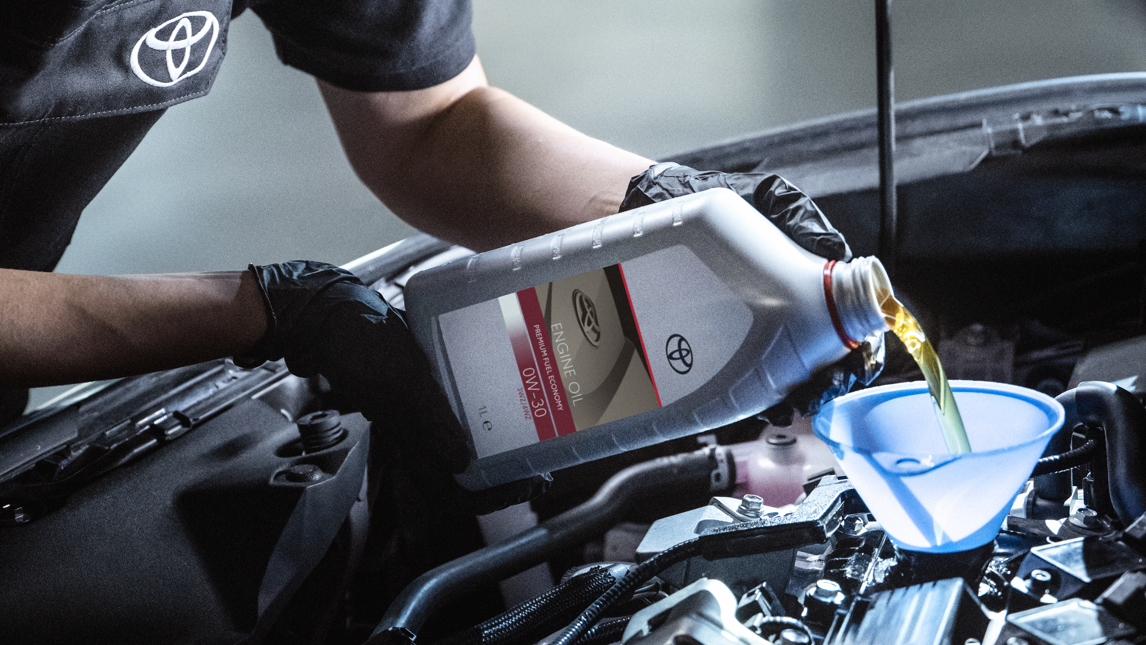Ein KFZ-Werkstattmitarbeiter in grauer Arbeitskleidung und Handschuhen gießt Öl in den Motor eines Toyota-Fahrzeugs.