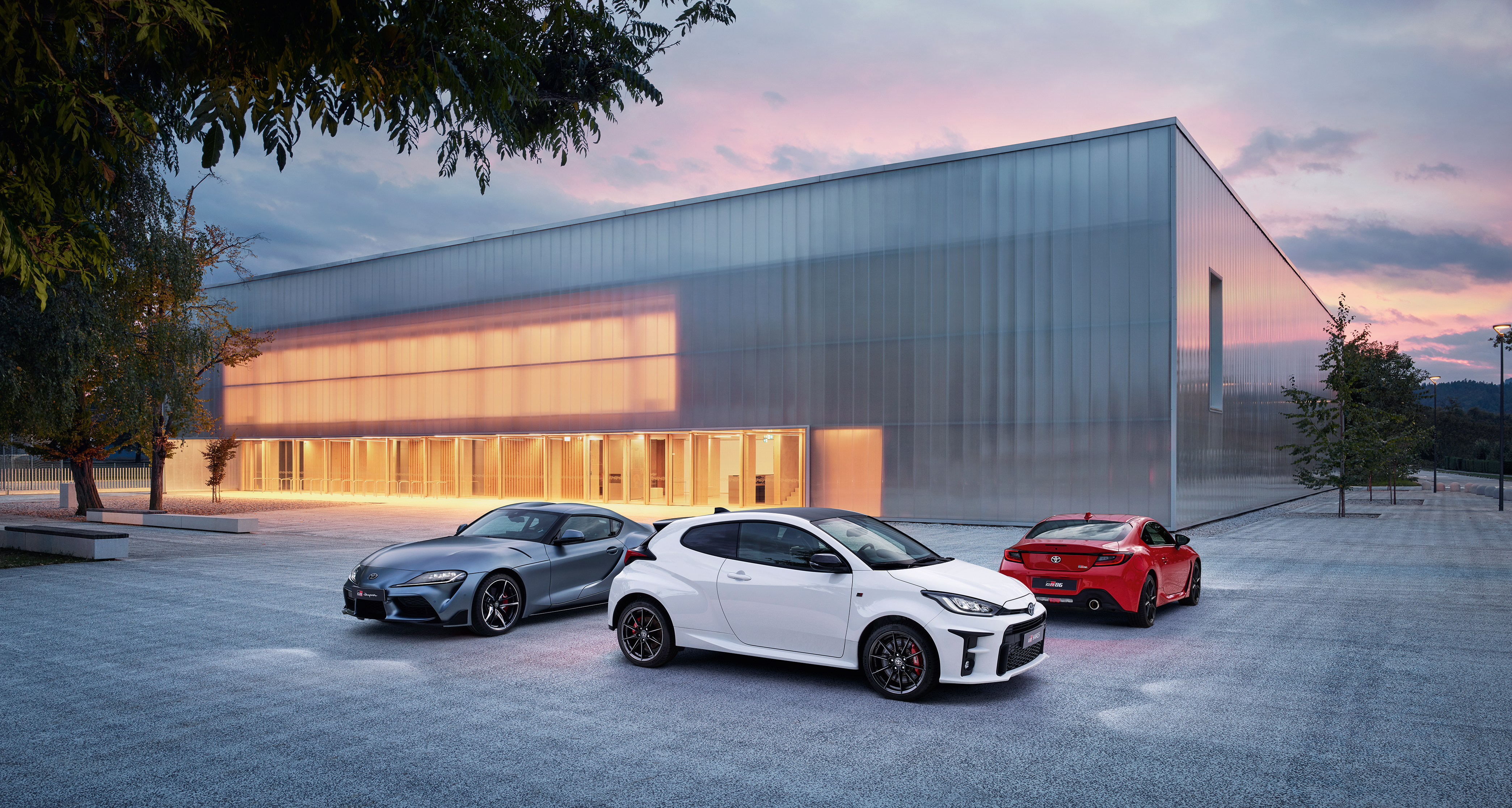 Drei GR Modelle stehen geparkt vor einer modernen Fabrikhalle. Ein weißer GR Yaris, ein roter GR 86 und ein grau-metallic-farbener GR Supra.