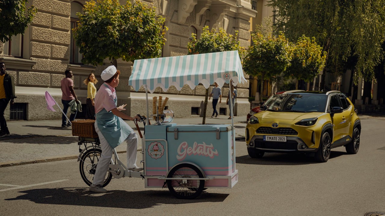 Ein rosa und hellblau gekleideter Eisverkäufer überquert mit seinem Eisverkauf-Fahrrad unerwartet vor einem Toyota Fahrzeug die Straße. Er gestikuliert dem Fahrer des Fahrzeugs anzuhalten. Passanten drehen sich erschrocken um.