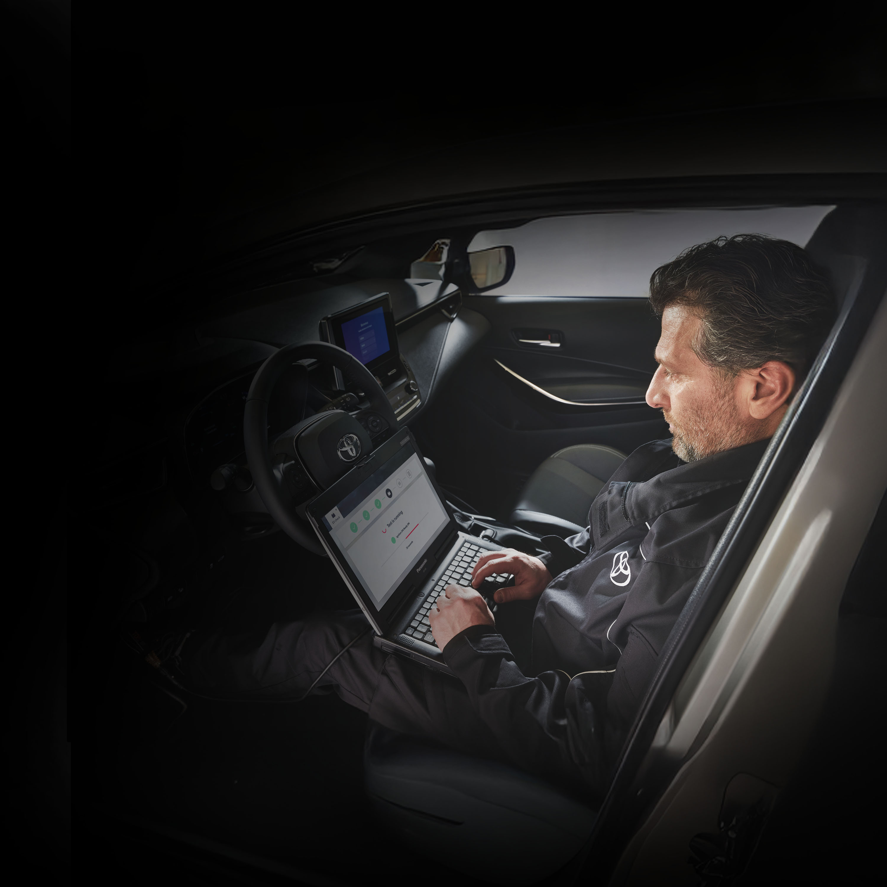 Ein bärtiger Mechaniker sitzt auf dem Fahrersitz eines Toyota und liest auf seinem Computer, den er auf dem Schoß hat, die Fehlermeldungen des Autos aus.