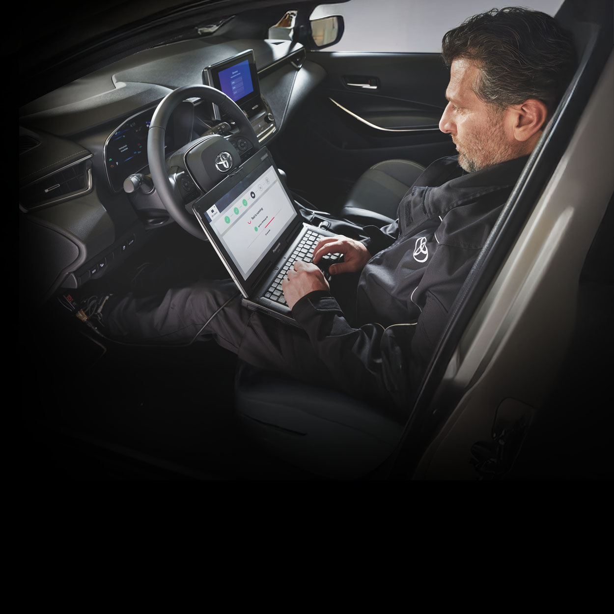Ein bärtiger Mechaniker sitzt auf dem Fahrersitz eines Toyota und liest auf seinem Computer, den er auf dem Schoß hat, die Fehlermeldungen des Autos aus.