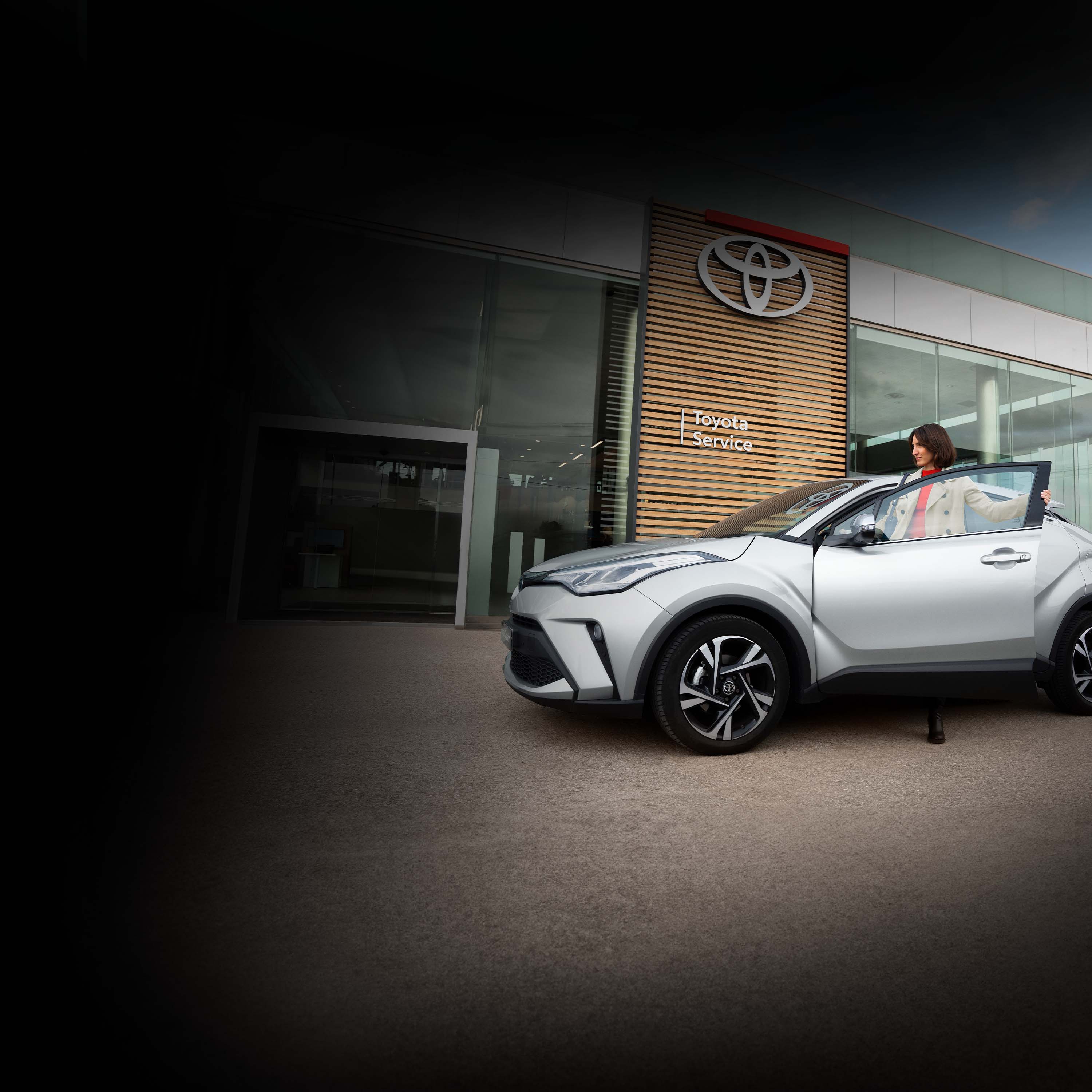 Eine lächelnde Frau steigt in den Fahrersitz eines silbernen Autos in Frontansicht. Hinter ihr ist der moderne, gläserene Eingang eines Autohauses, an dessen Wand die Aufschrift Toyota Service steht.