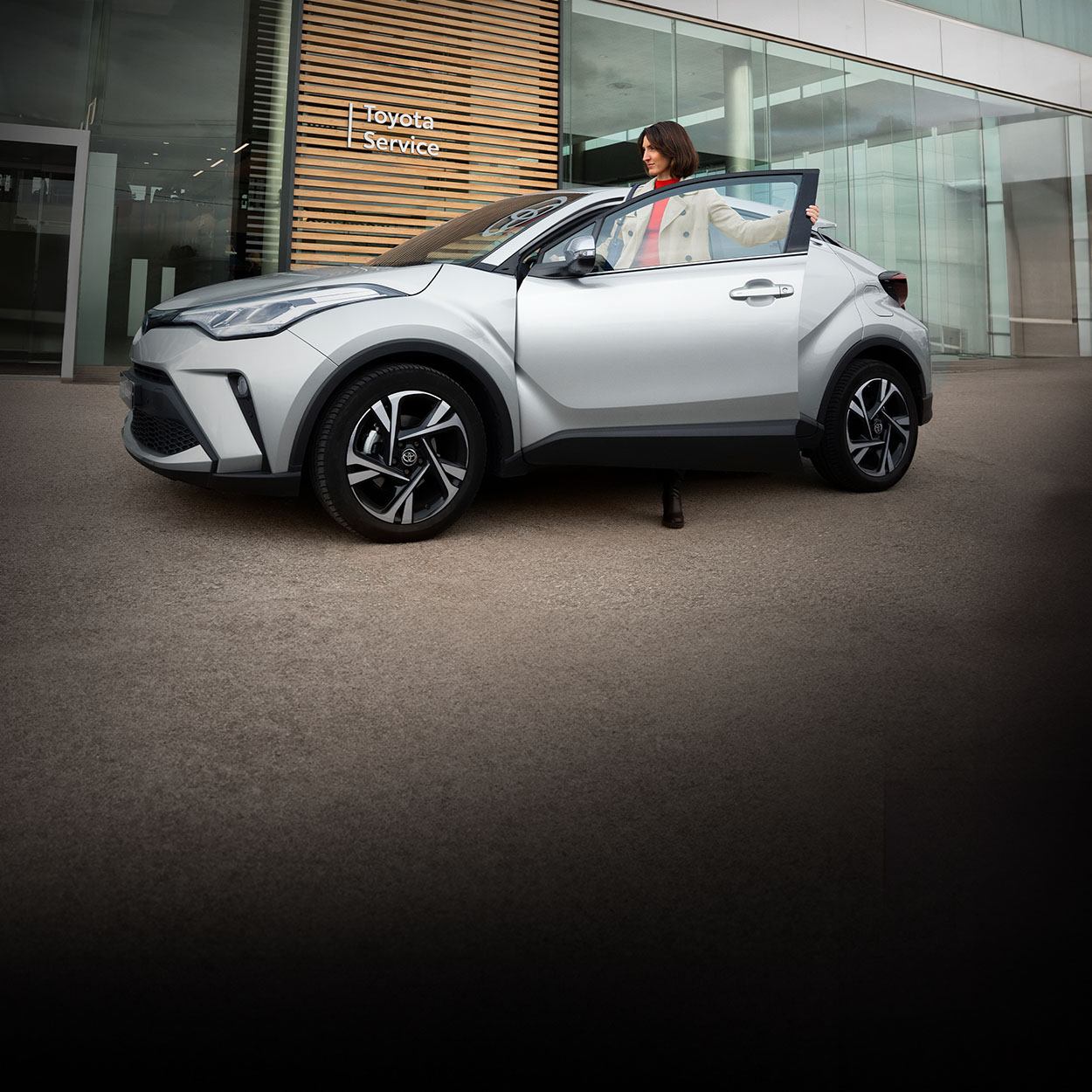 Eine lächelnde Frau steigt in den Fahrersitz eines silbernen Autos in Frontansicht. Hinter ihr ist der moderne, gläserene Eingang eines Autohauses, an dessen Wand die Aufschrift Toyota Service steht.