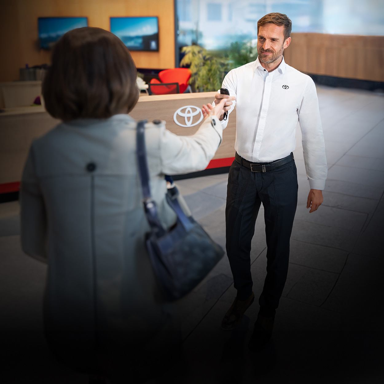 Ein lächelner Mann, der ein weißes Hemd mit dem Toyota-Logo trägt, reicht einer Dame mit dunklen Haaren einen Autoschlüssel in die ausgestreckte Hand. Im Hintergrund erkennt man eine Rezeption.