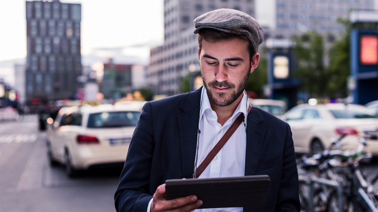 Ein Mann mit grauer Schiebermütze, weißem Hemd und schwarzem Sakko schaut auf ein Tablet. Im Hintergrund ein Taxistand.