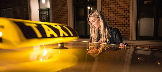 Im Vordergrund das Dach eines Taxis mit einem leuchtenden Taxi-Schild. Eine Frau steigt in das Taxi ein.