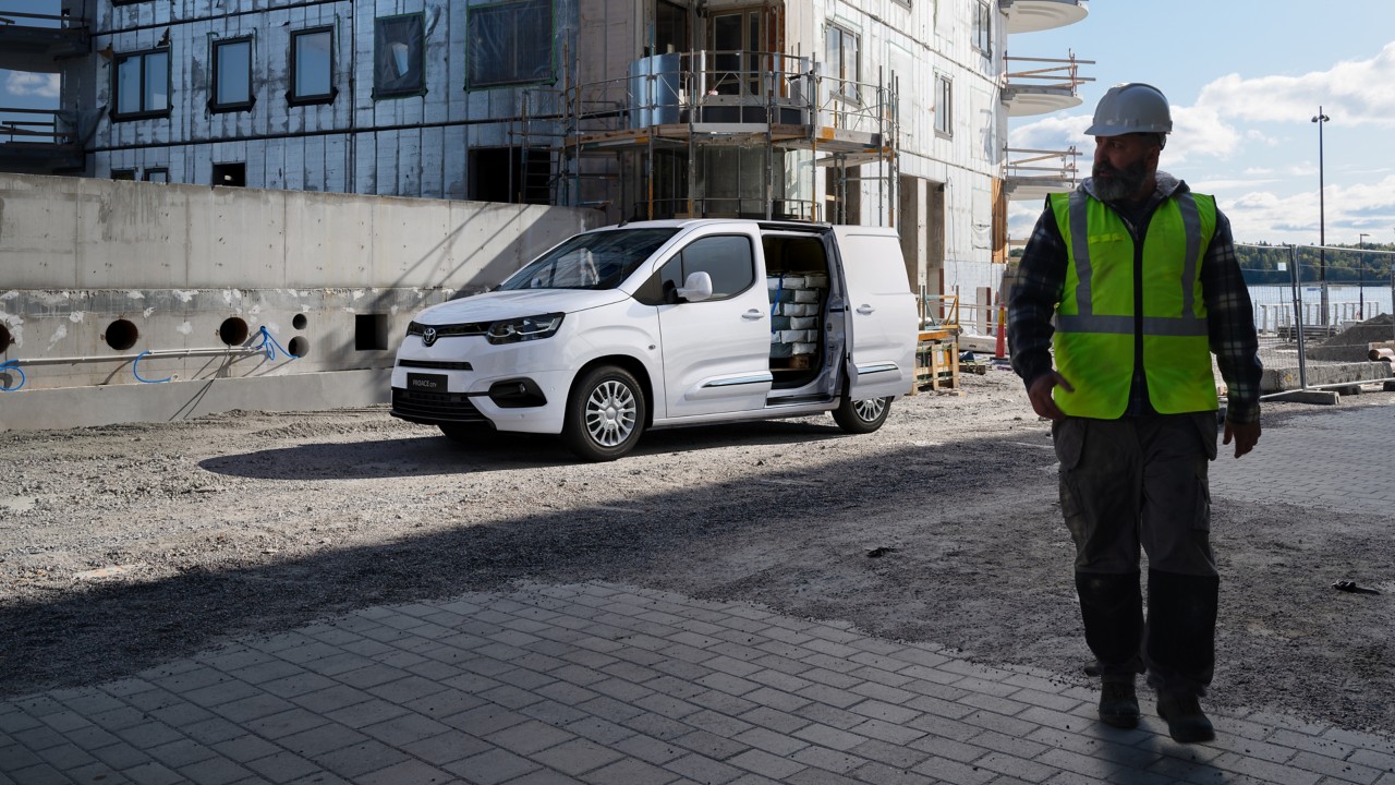 Ein Bauarbeiter mit weißem helm und neon-gelber Sicherheitsweste steht im Vordergrund. Hinter ihm eine Baustelle mit einem beladenen Proace City.
