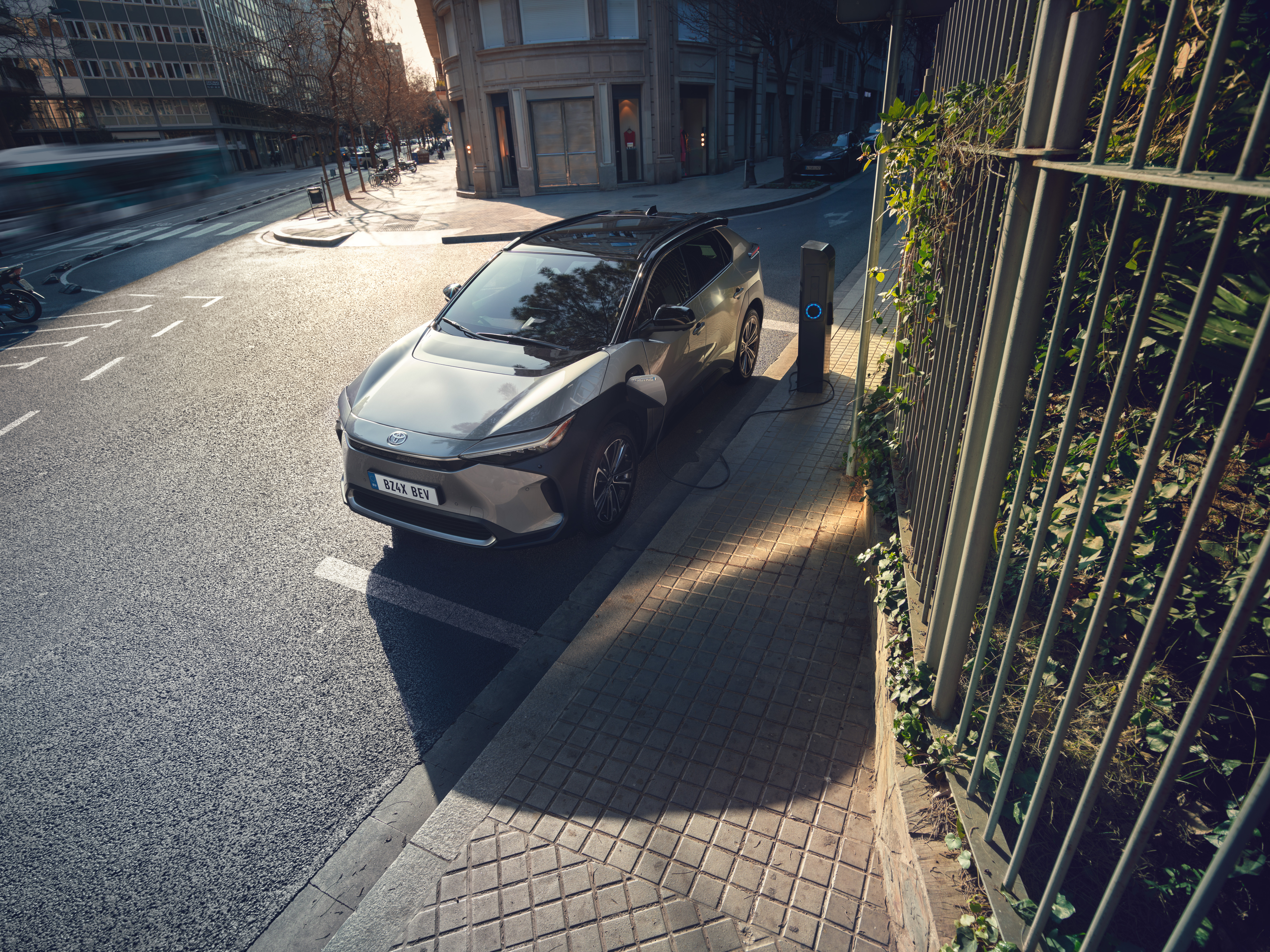 Toyota BZ4X steht an einer öffentlichen Ladesäule am Straßenrand in einer urbanen Umgebung und wird bei Tageslicht aufgeladen.