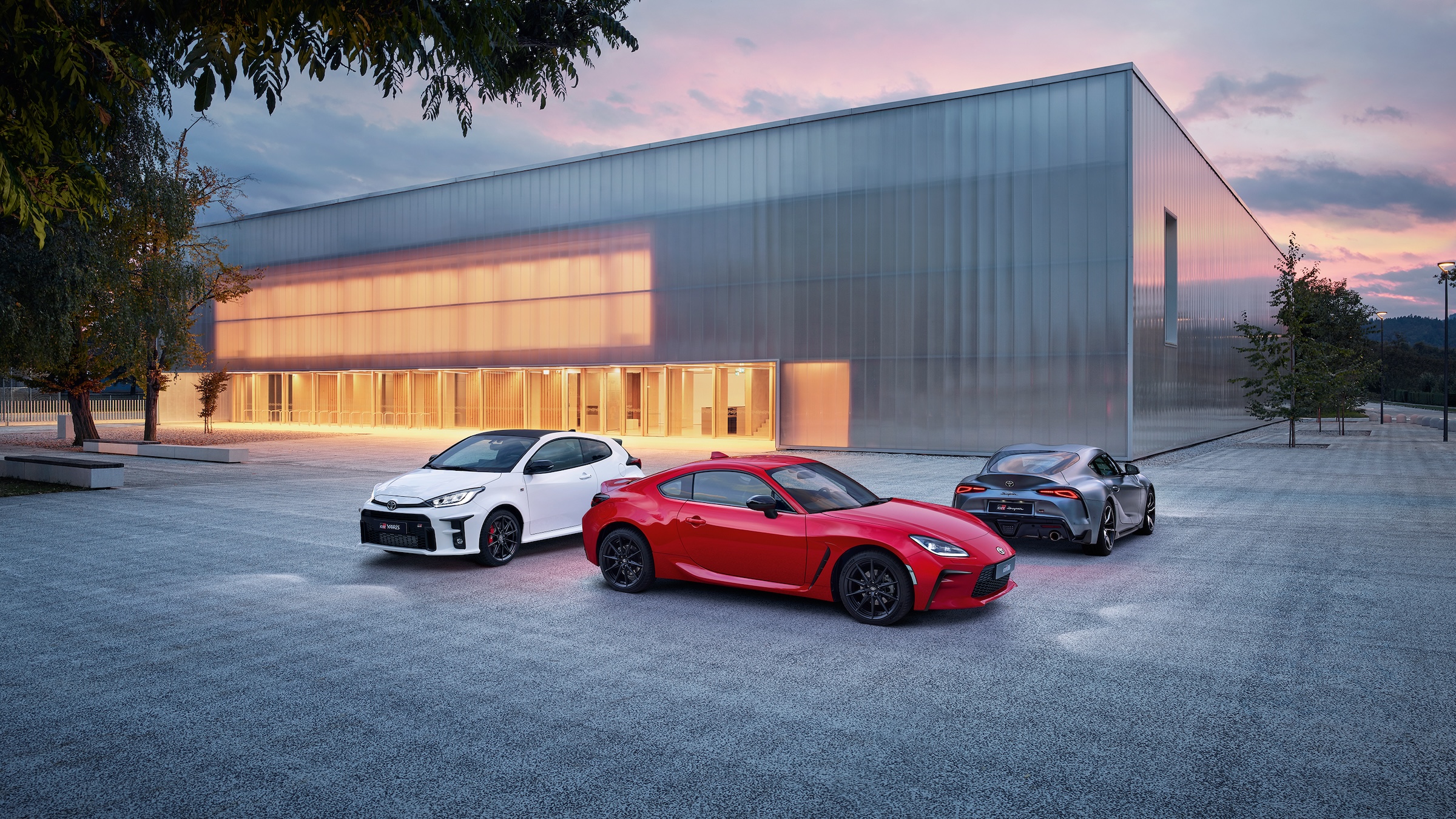 Ein GR Yaris, GR86 und GR Supra in der Dämmerung, geparkt vor einer modernen Veranstaltungshalle mit erleuchtetem Eingangsbereich