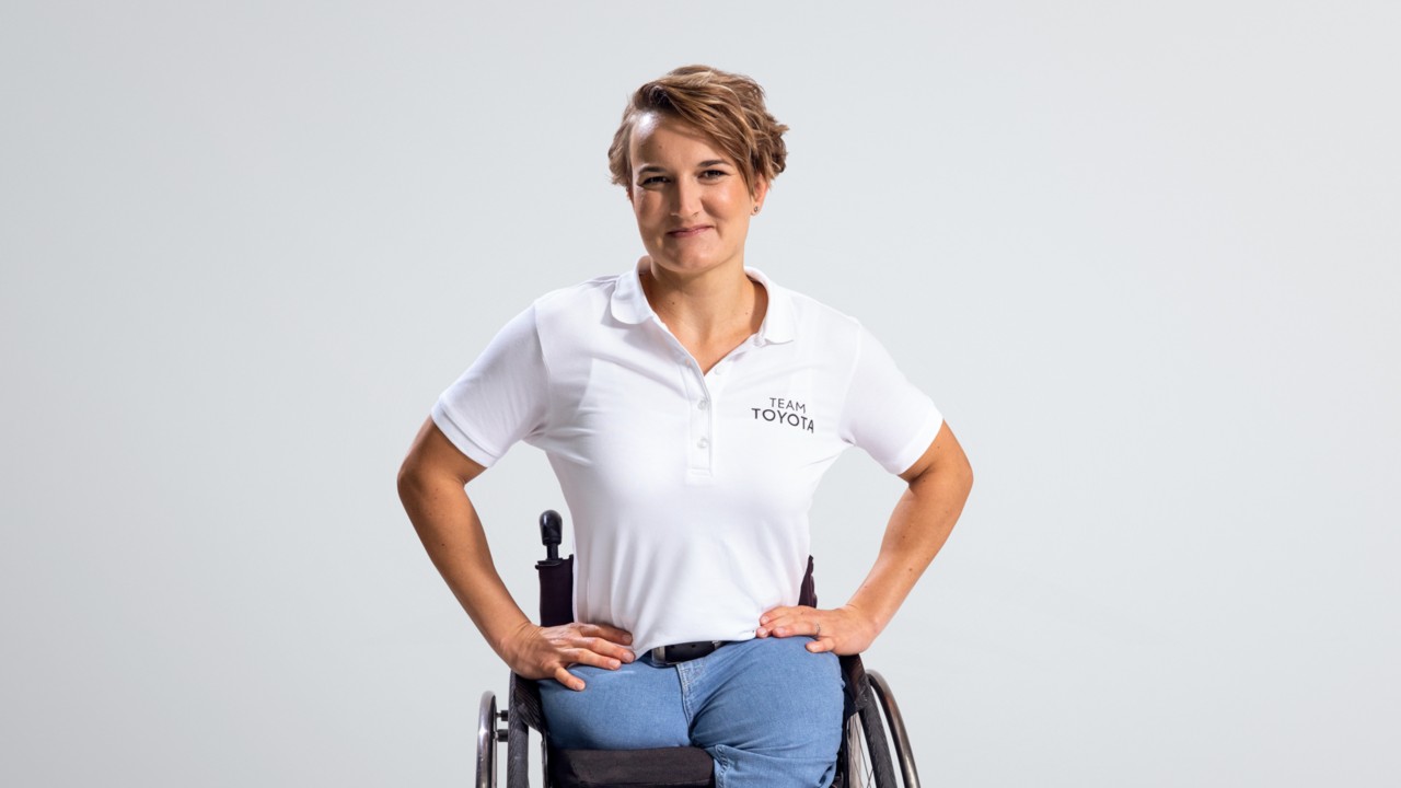Eine junge Frau im Rollstuhl mit weißem Shirt und Jeans lächelt in die Kamera: Anna-Lena Foster. Ab der Hüfte fehlen ihr die Beine. 