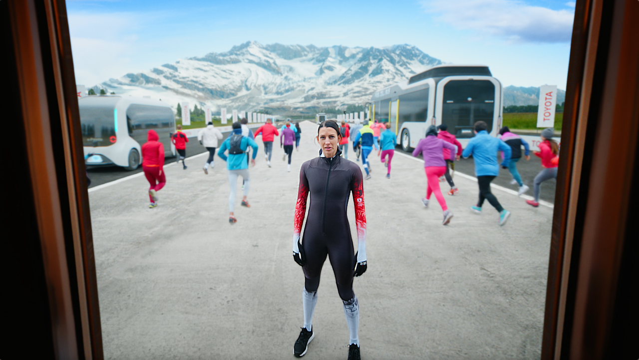 Eine Frau im Sportleroutfit steht zur Kamera zugewandt. Dahinter viele Menschen die in richtung Horizont laufen, wo eine Berglandschaft zu sehen ist.