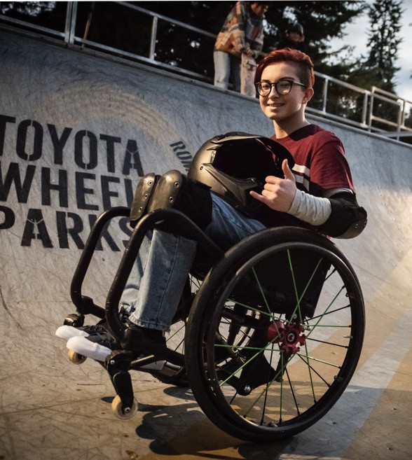 Eine junge Frau im Rollstuhl und Motorradhelm auf dem Schoß, Lilly Rice, macht einen Wheelie auf der Halfpipe und lächelt in die Kamera