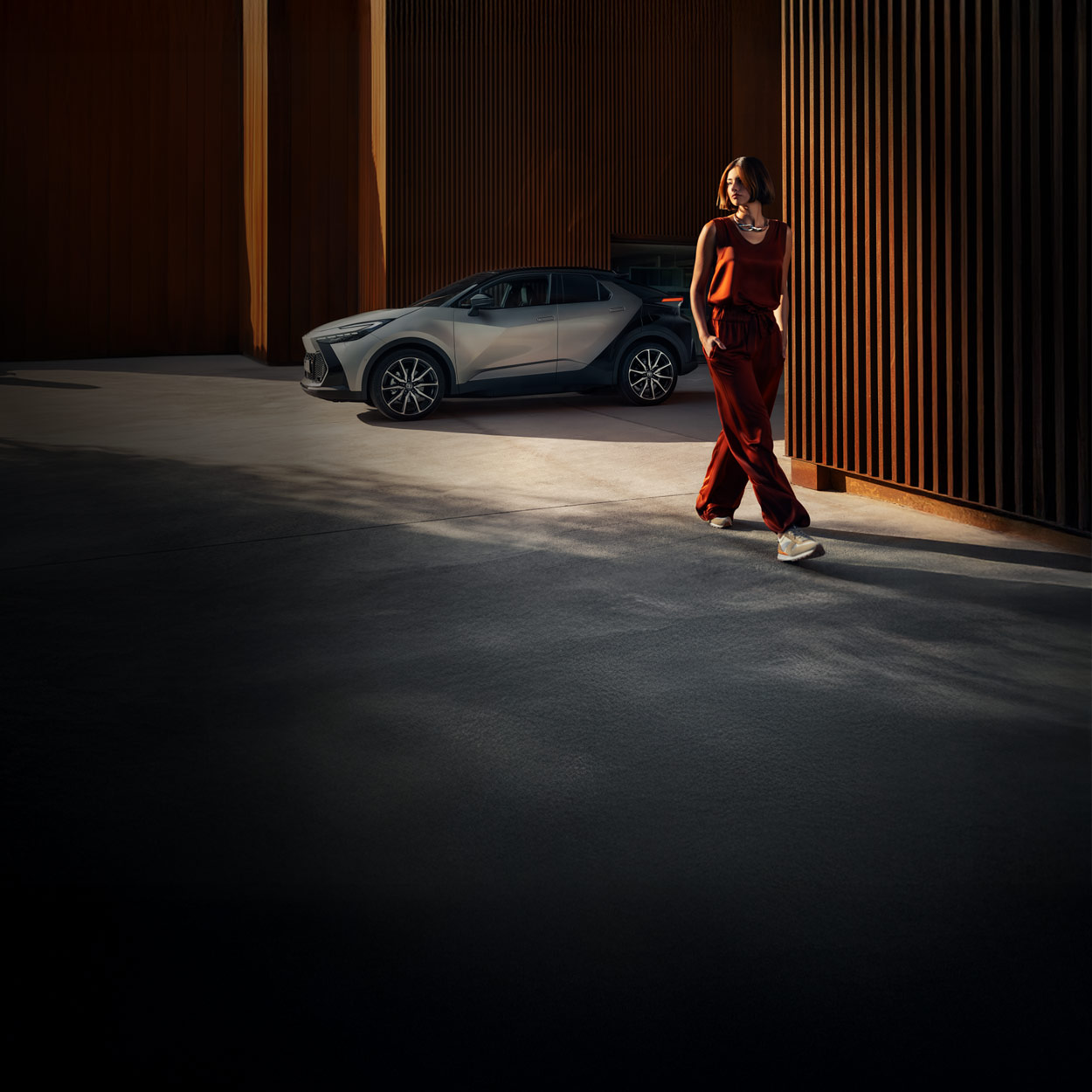 Eine Frau in einem kastanienbraunen Outfit geht durch eine moderne, industrielle Umgebung im Freien mit einem silbernes Auto, das in der Nähe geparkt ist.